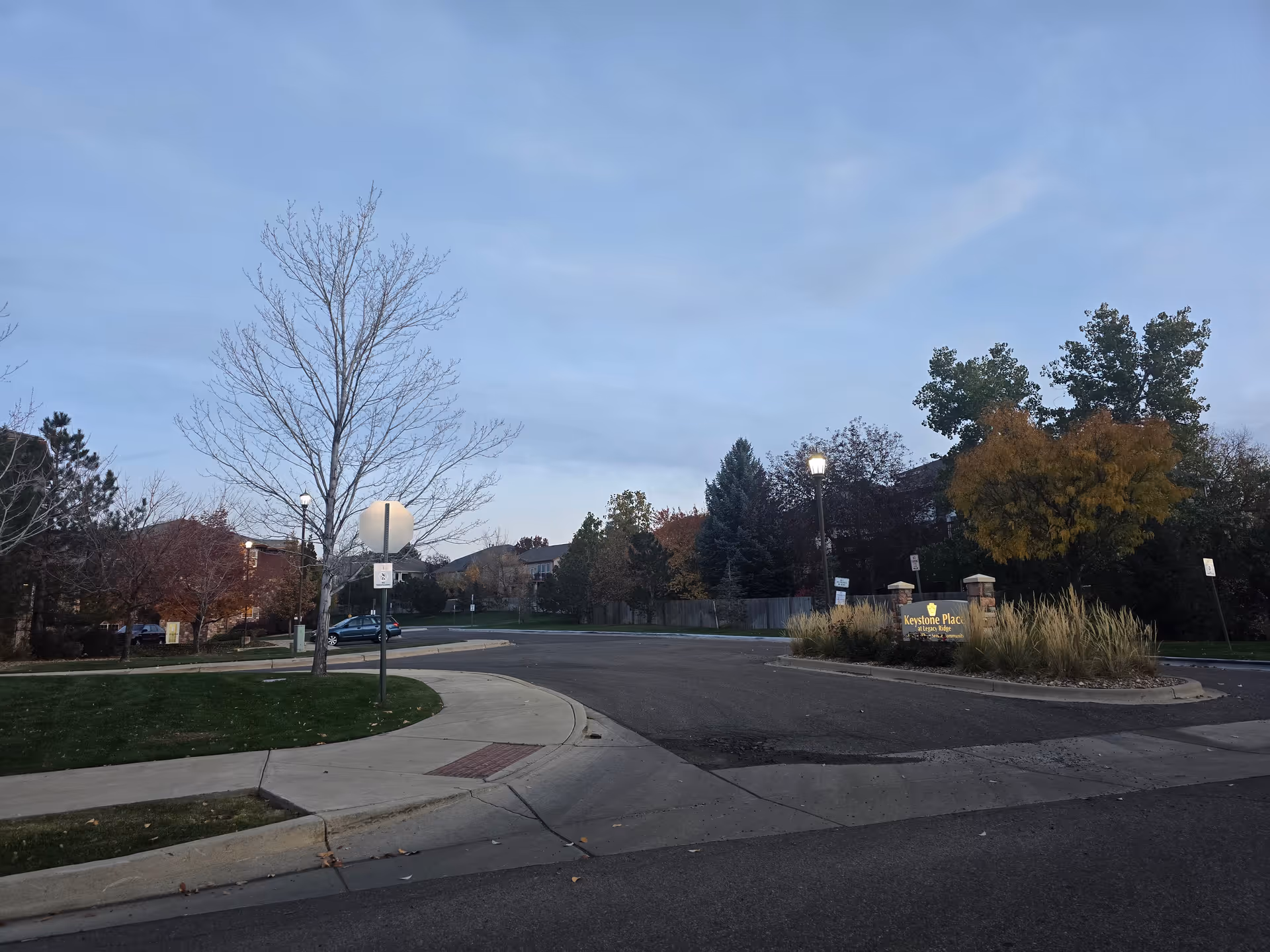 Street entrance and landscaped roundabout with a Keystone Place sign in front of trees and buildings at dusk.