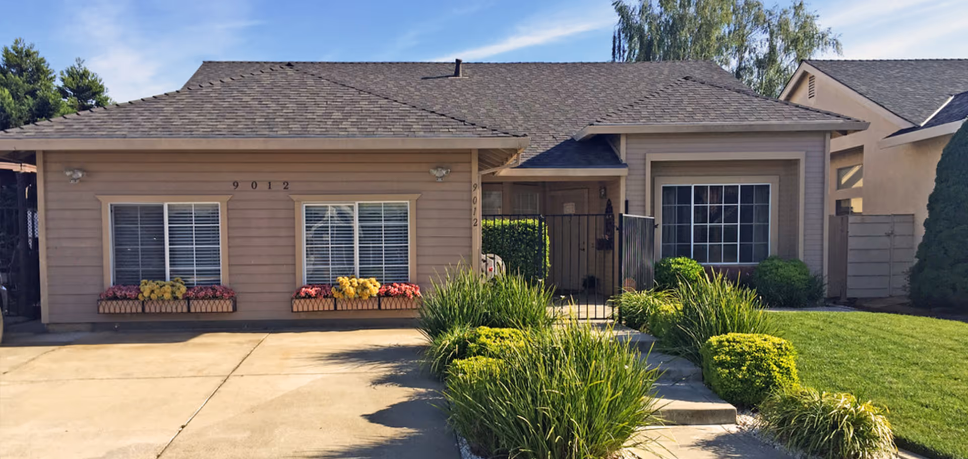 Single-story beige house front with two large windows, planter boxes, a gated entryway, and a neatly landscaped lawn and driveway.