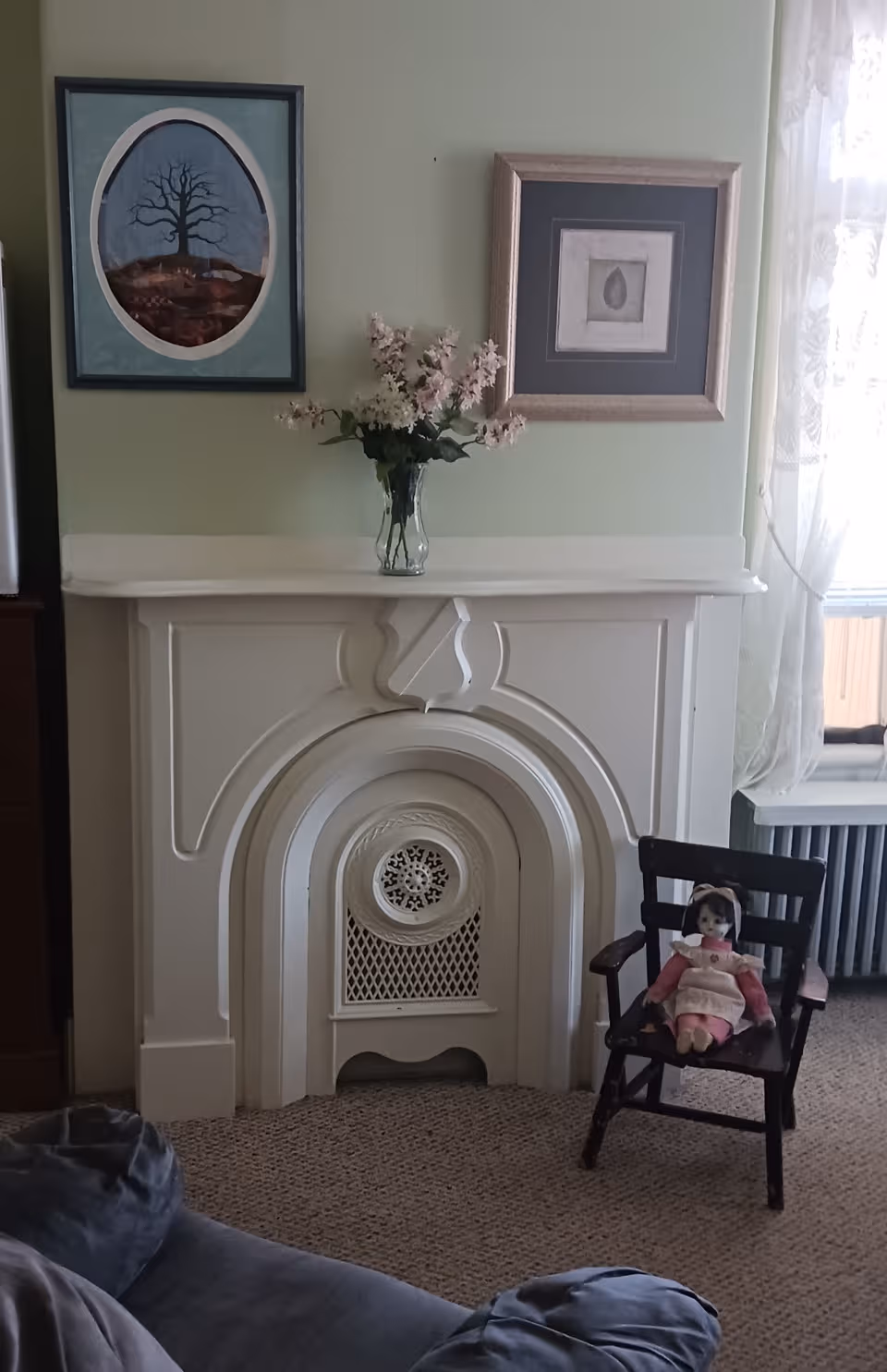Pastel living room with a white fireplace, framed art, flowers, and a doll on a chair.