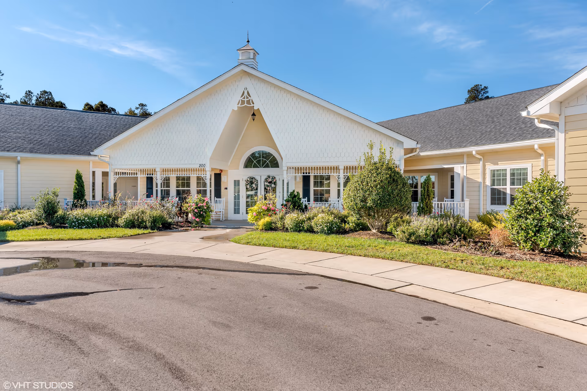 Front exterior view of a single-story senior living facility building with a peaked roof, white trim, and beige siding. The entrance features a covered porch with white rocking chairs and is surrounded by well-maintained landscaping including bushes and flowers. The sky is clear and blue.
