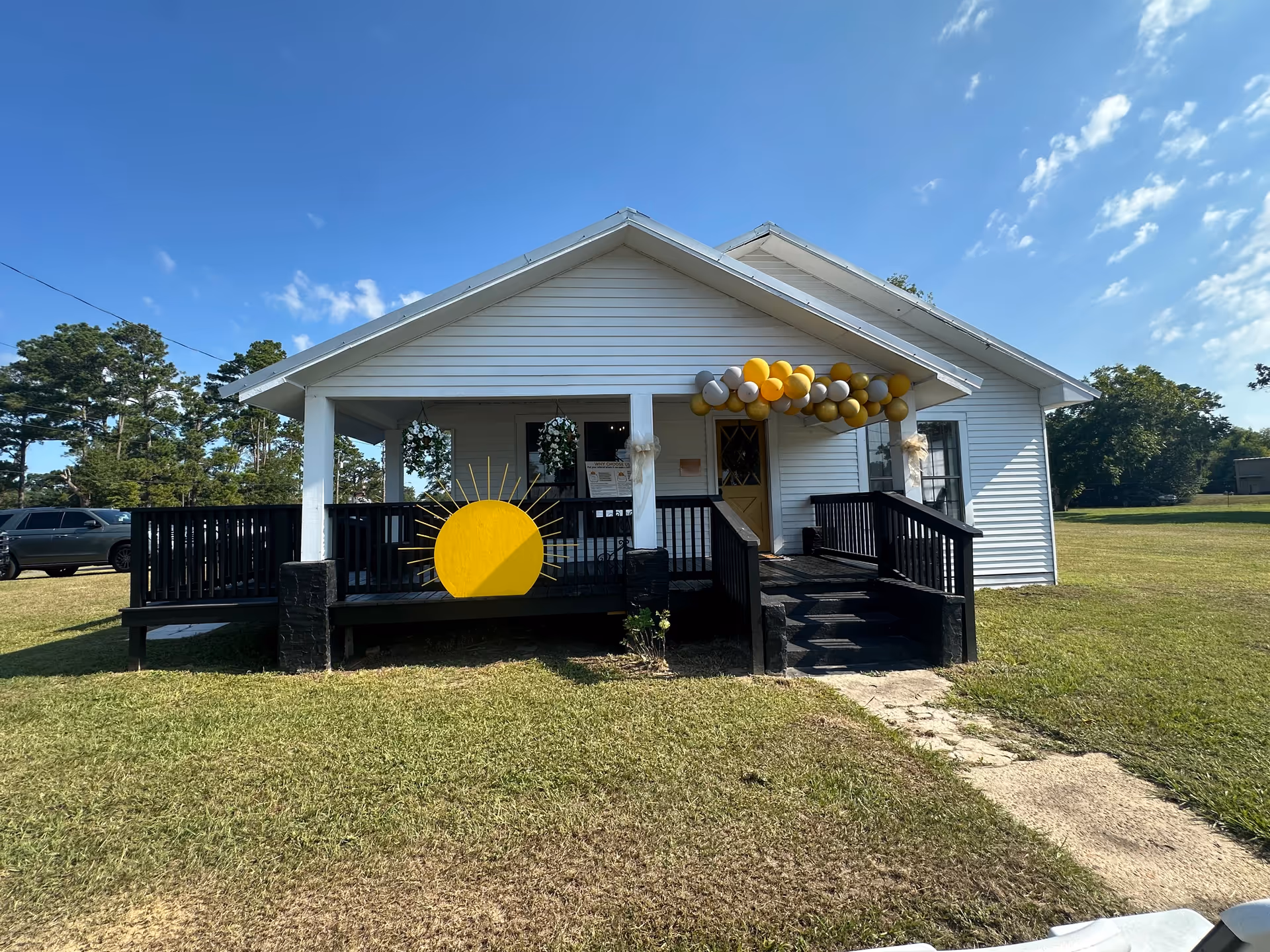 White one-story building with a covered front porch, balloons, and a yellow sun decoration outside.