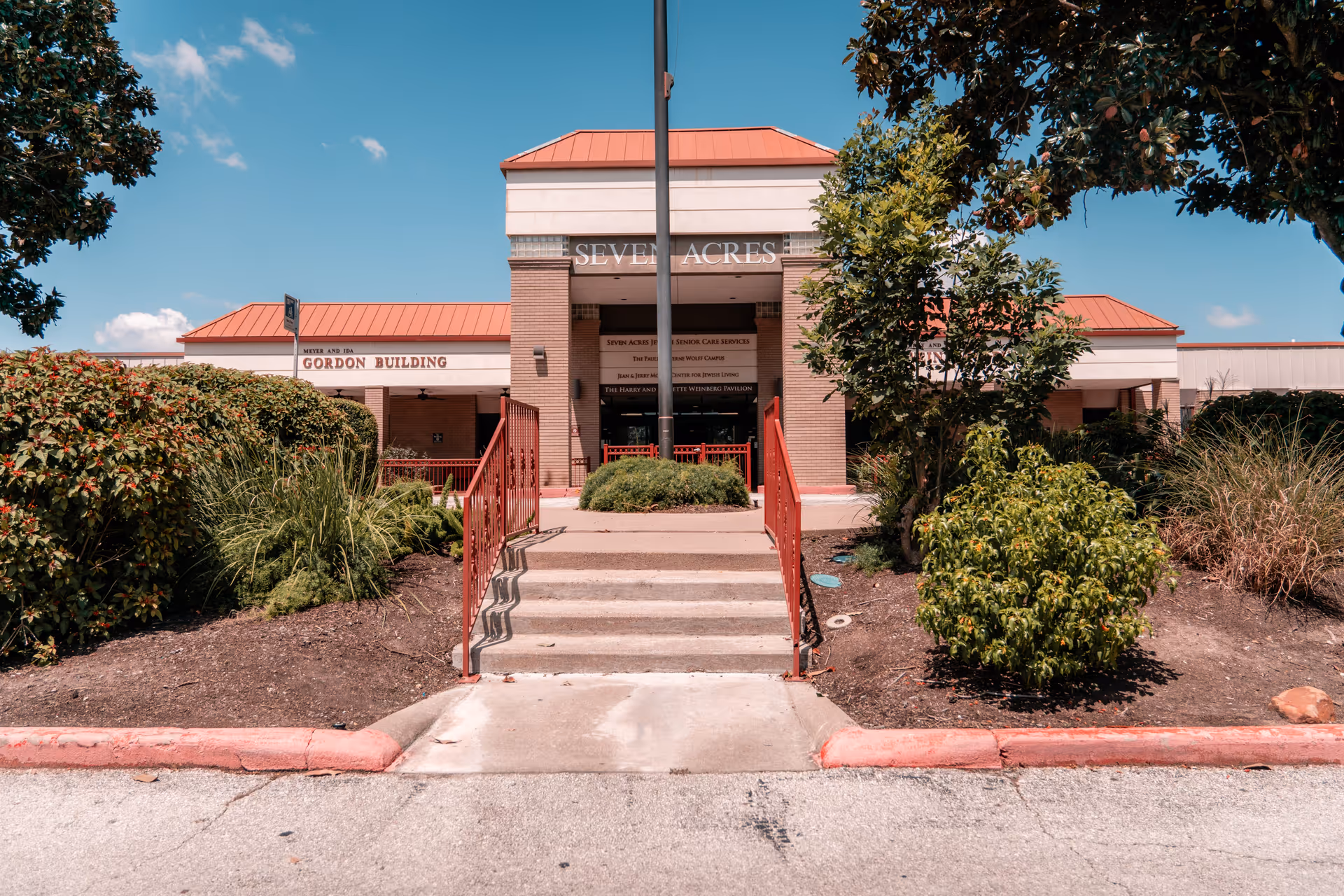 Front entrance of the Seven Acres senior care building with steps, red railings, and landscaped shrubs under a blue sky.