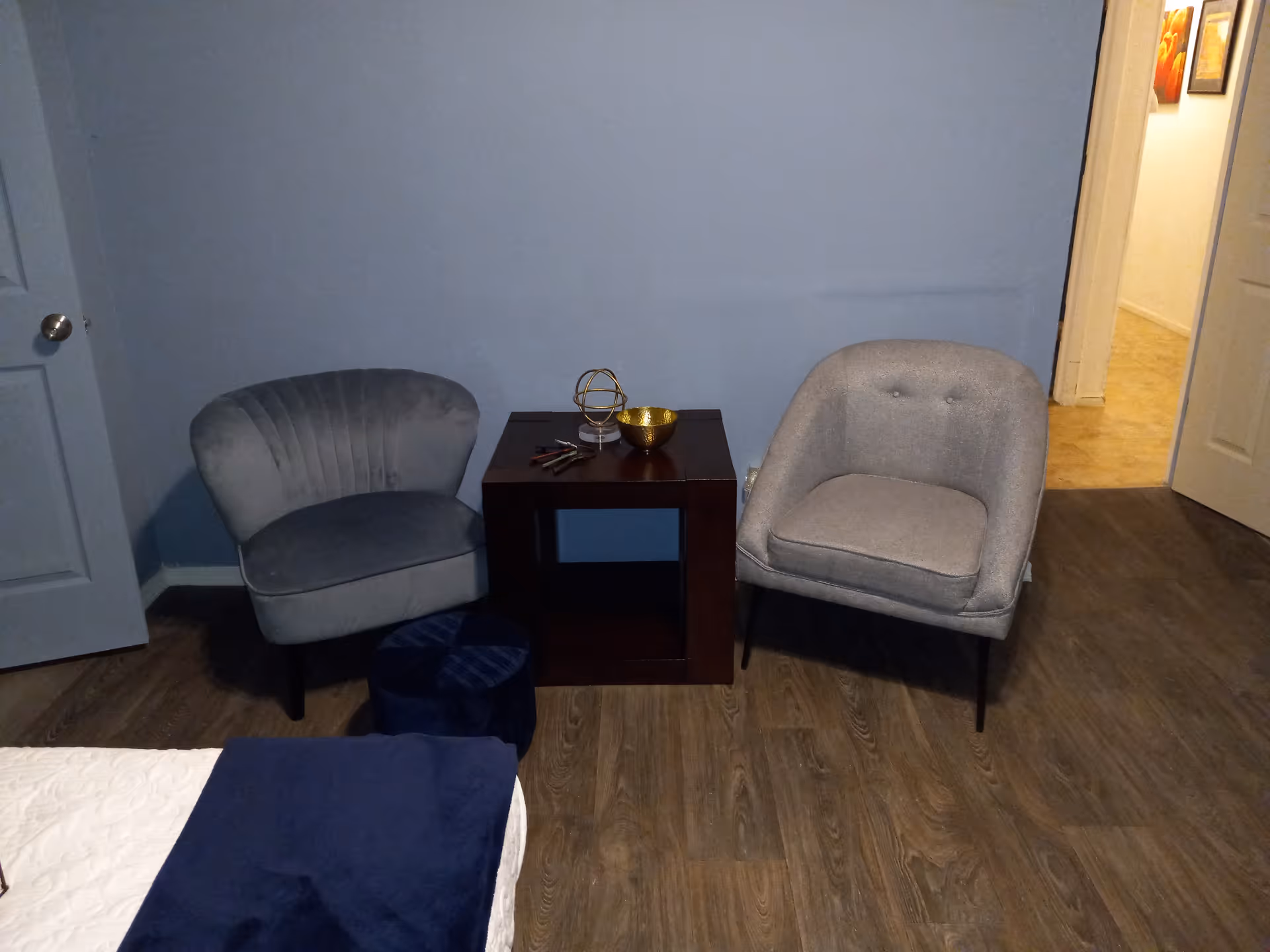 Two gray chairs flank a dark wooden side table in a small blue-walled living area.