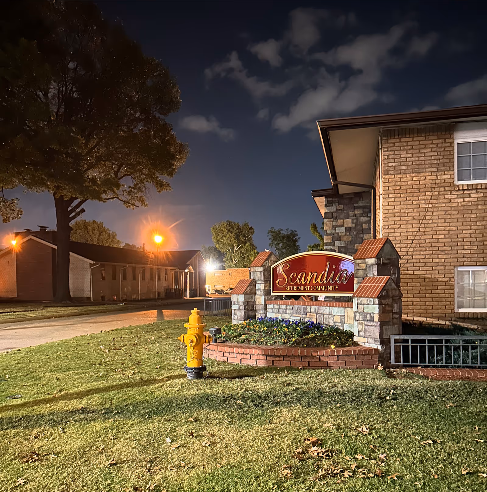 Night exterior view of Scandia Retirement Community sign beside brick building and landscaped garden.