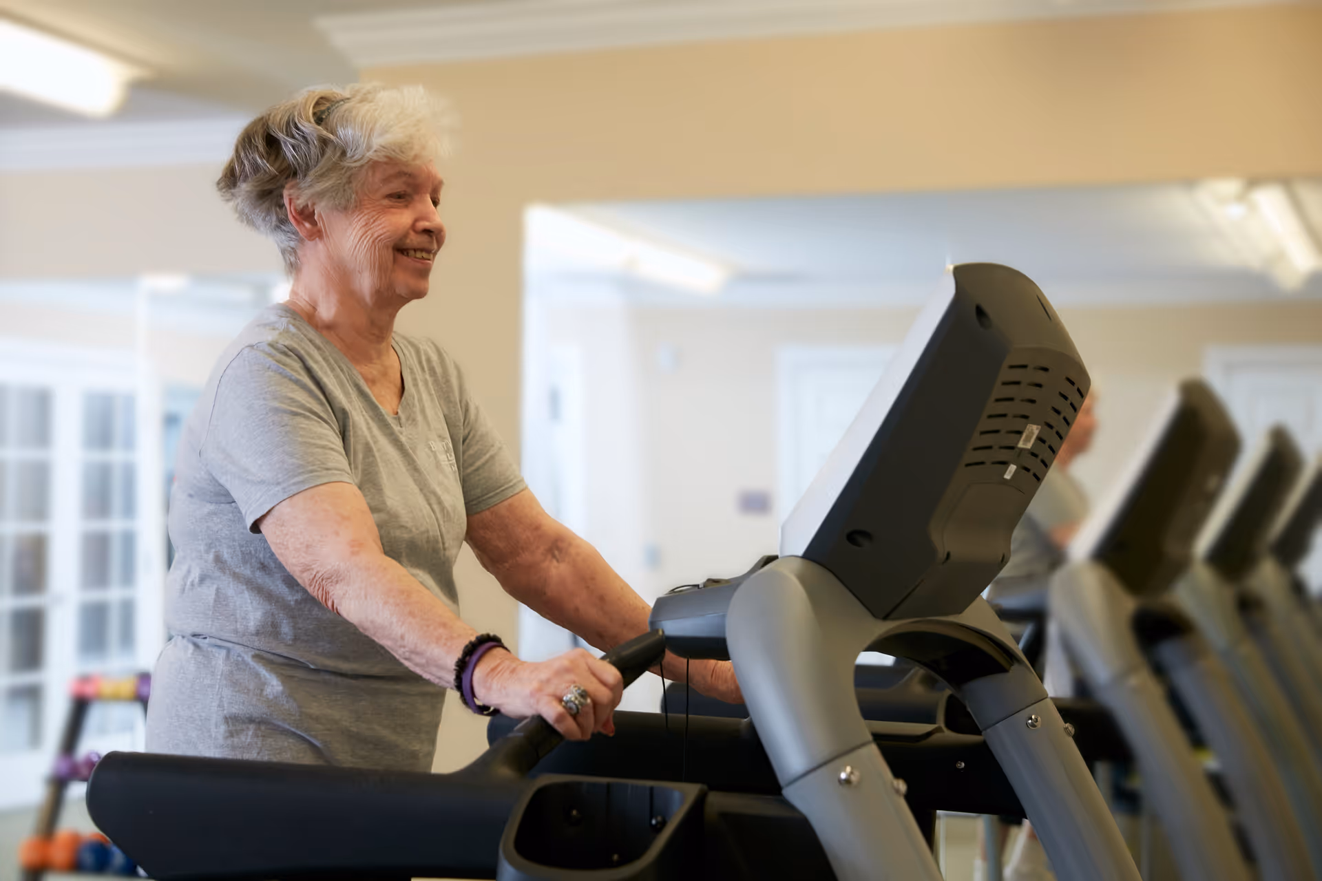 An elderly woman with short gray hair is smiling while walking on a treadmill in a fitness room with mirrors and bright lighting.