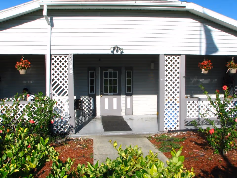 Front entrance of a single-story building with white siding and a covered porch. The porch has white lattice panels and hanging flower pots with red flowers. There is a concrete walkway leading to a door with glass panels, flanked by two narrow windows. Green shrubs and plants are visible in the foreground.