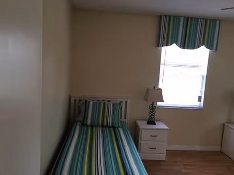 Bedroom at Acacia Grove Assisted Living Facility LLC with striped bed, nightstand, lamp, and window.
