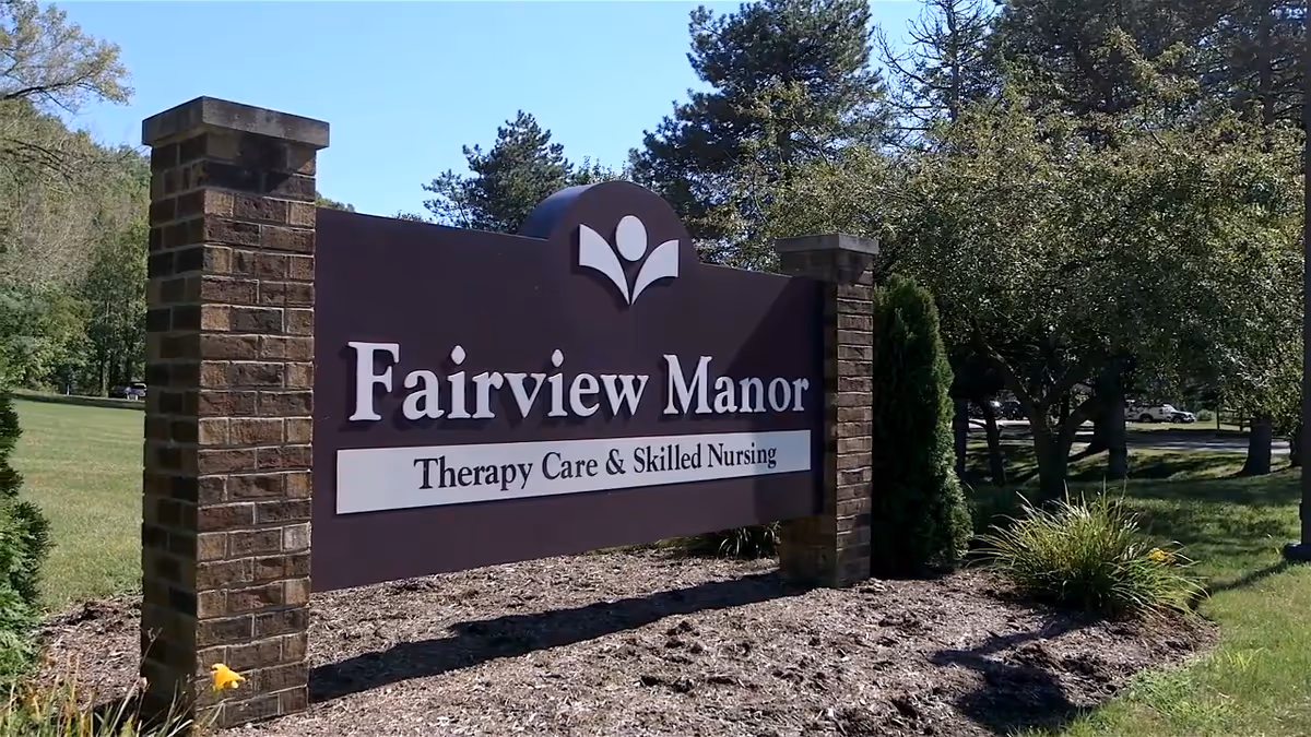 Outdoor sign reading Fairview Manor Therapy Care and Skilled Nursing amid trees and shrubs.