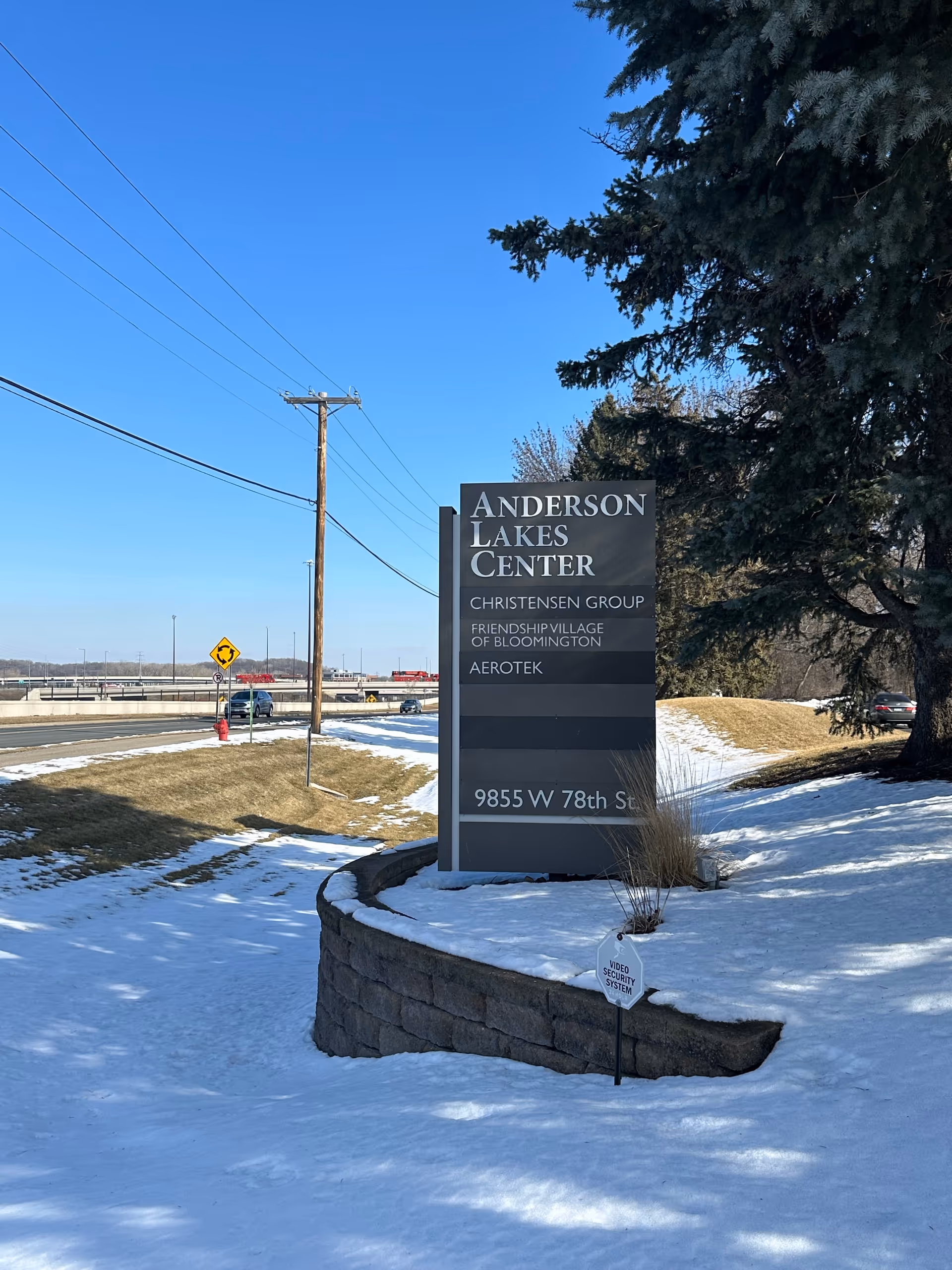 Snowy outdoor sign for Anderson Lakes Center beside road and trees under blue sky