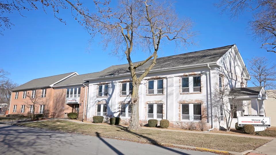 Two-story brick-and-white facility exterior with trees and a sidewalk.