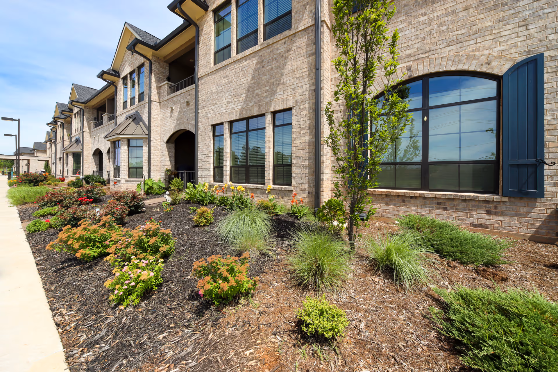 Landscaped brick building front with large arched windows, shrubs, and a sidewalk.