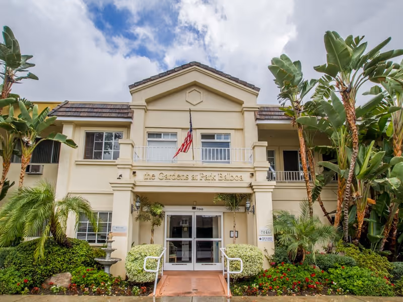 Front exterior of The Gardens at Park Balboa with entry doors, flag, and tropical landscaping.