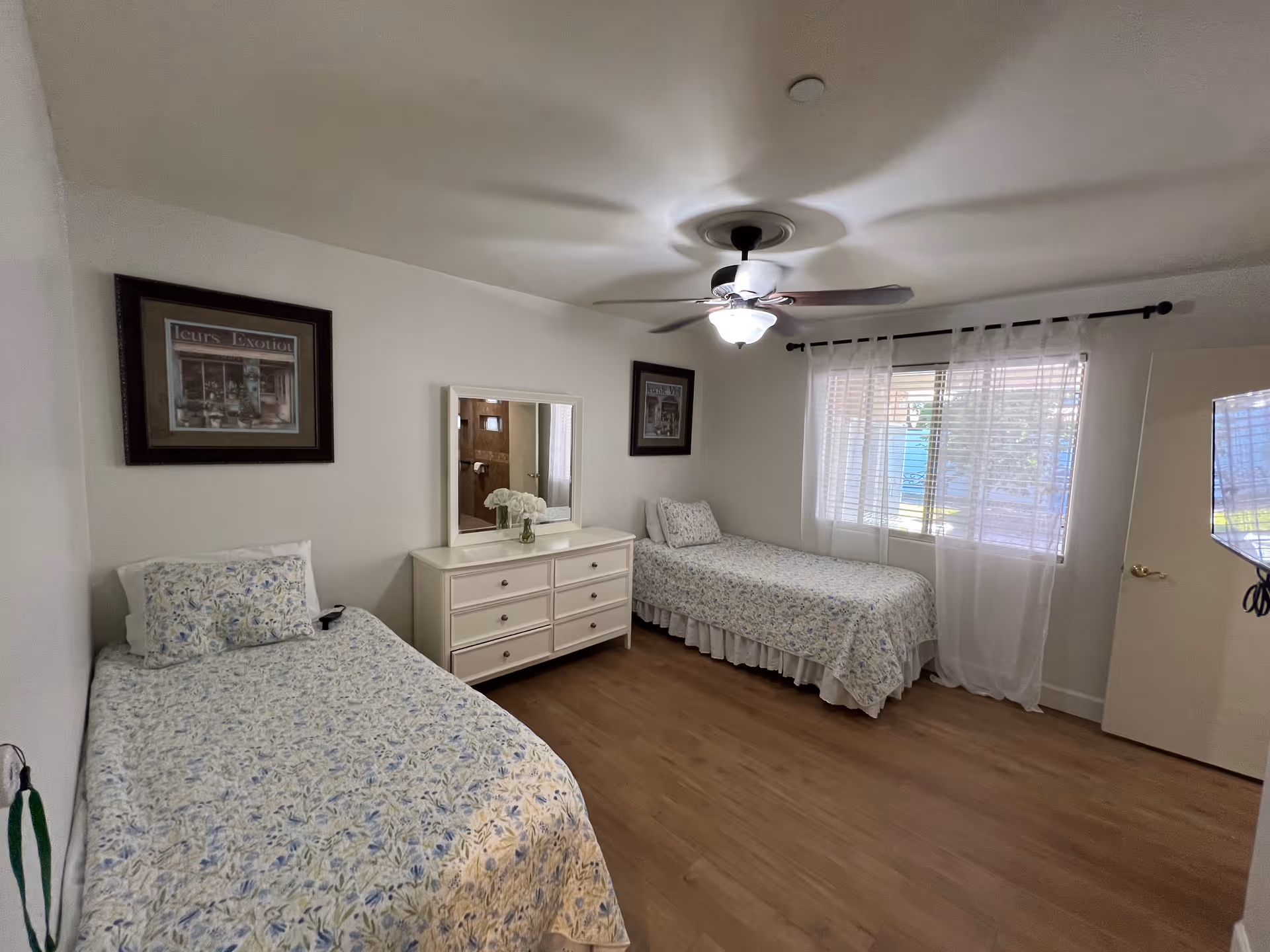 A bright bedroom with two single beds covered in floral bedspreads, a white dresser with a mirror and a vase of flowers, a ceiling fan with light, a window with sheer white curtains, and two framed pictures on the walls.