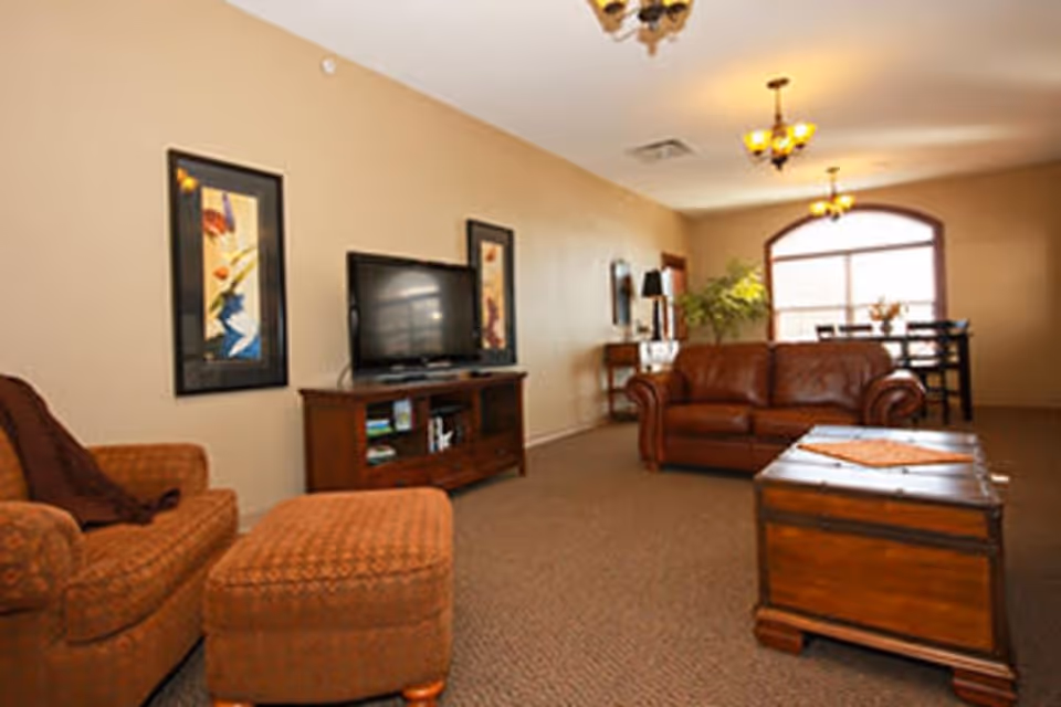 A cozy living room with beige walls and carpeted floor featuring a brown leather sofa, an upholstered armchair with matching ottoman, a wooden TV stand with a flat-screen television, two framed abstract paintings on the wall, a wooden chest used as a coffee table, and a dining area with a table and chairs near a large arched window letting in natural light.