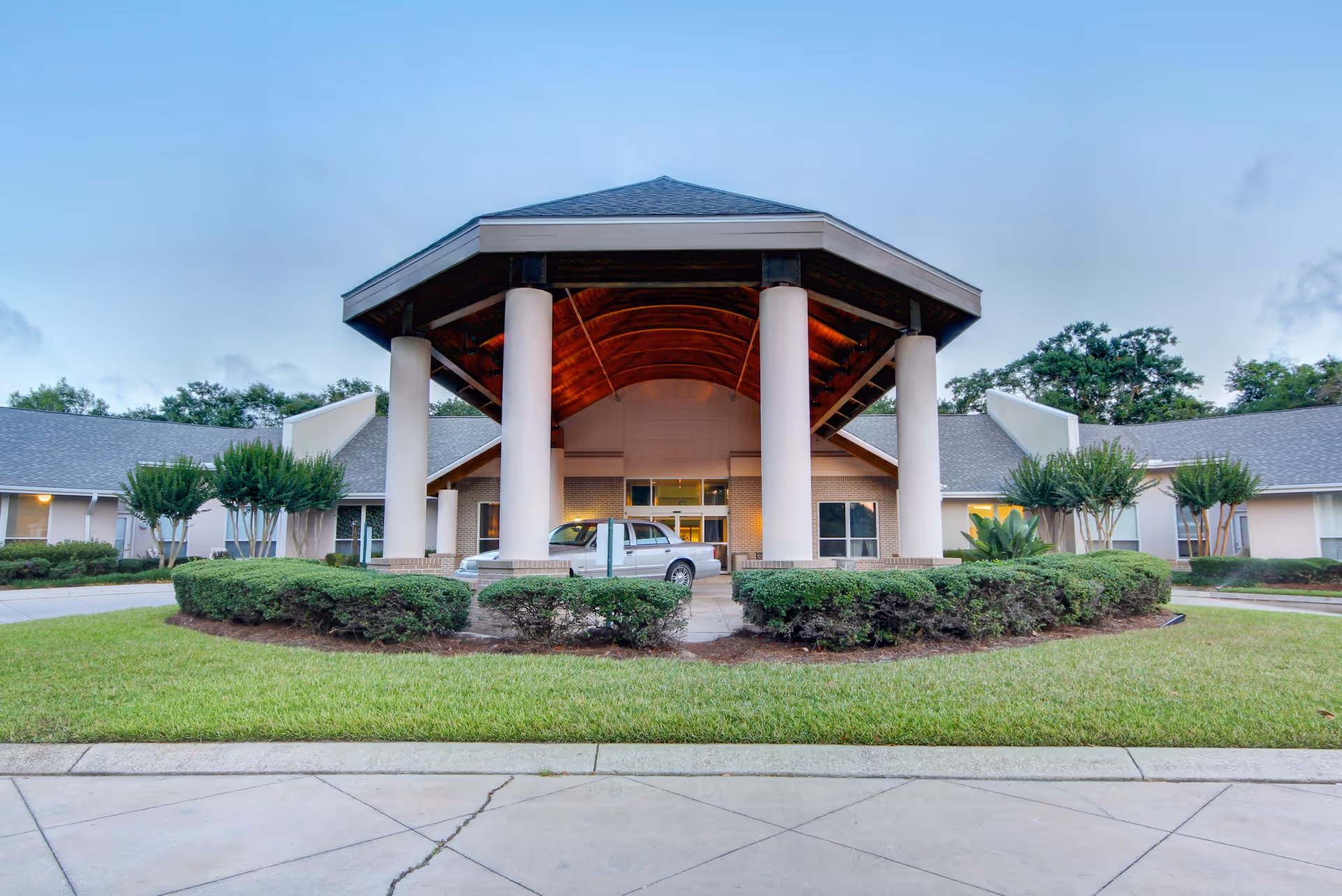 Front entrance of a senior living building with a large covered porte-cochère supported by columns, a parked car, and landscaped shrubs and lawn.