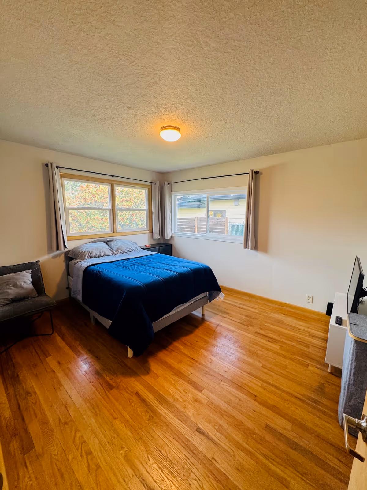 A tidy bedroom with a bed, a chair, wooden floor, and two windows.