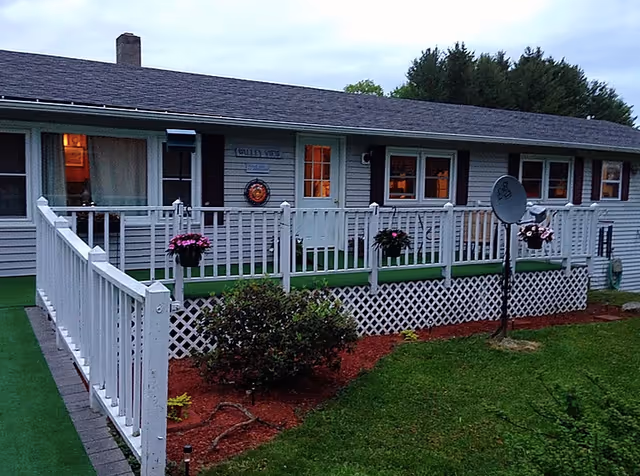 Exterior view of a single-story building with a white railing and a small porch area decorated with hanging flower pots. The building has several windows and a door, with a satellite dish mounted on a pole in the front yard. The lawn is green and well-maintained with some bushes and mulch beds.