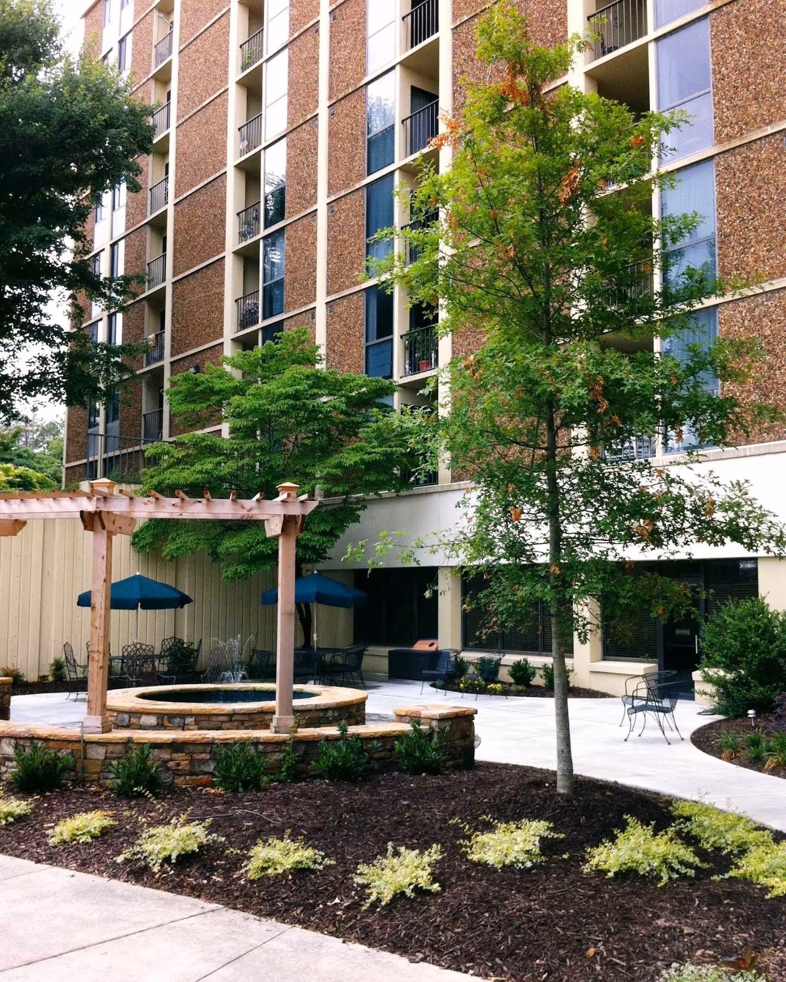 Outdoor courtyard area of a multi-story building with a stone fountain surrounded by a wooden pergola, green trees, shrubs, and patio seating with tables and umbrellas.