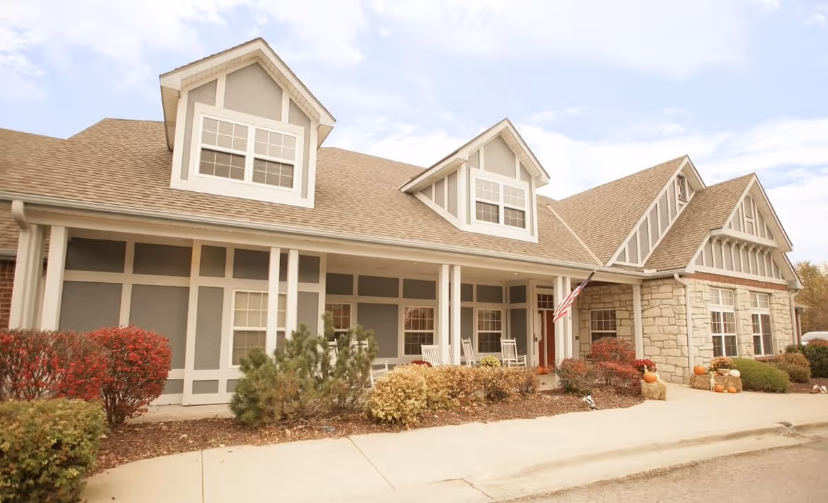 Exterior view of Homestead Estates Assisted Living of Leawood showing a single-story building with a beige and gray facade, multiple windows, a covered porch with white rocking chairs, an American flag, and landscaping with bushes and pumpkins near the entrance.