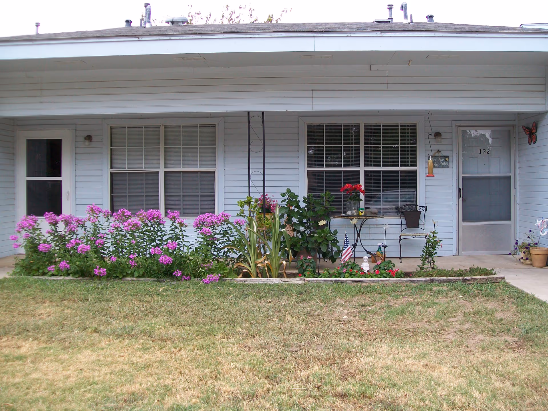 Single-story building front with a covered porch, windows and doors, a small patio table and chairs, and a flowerbed of purple blooms.