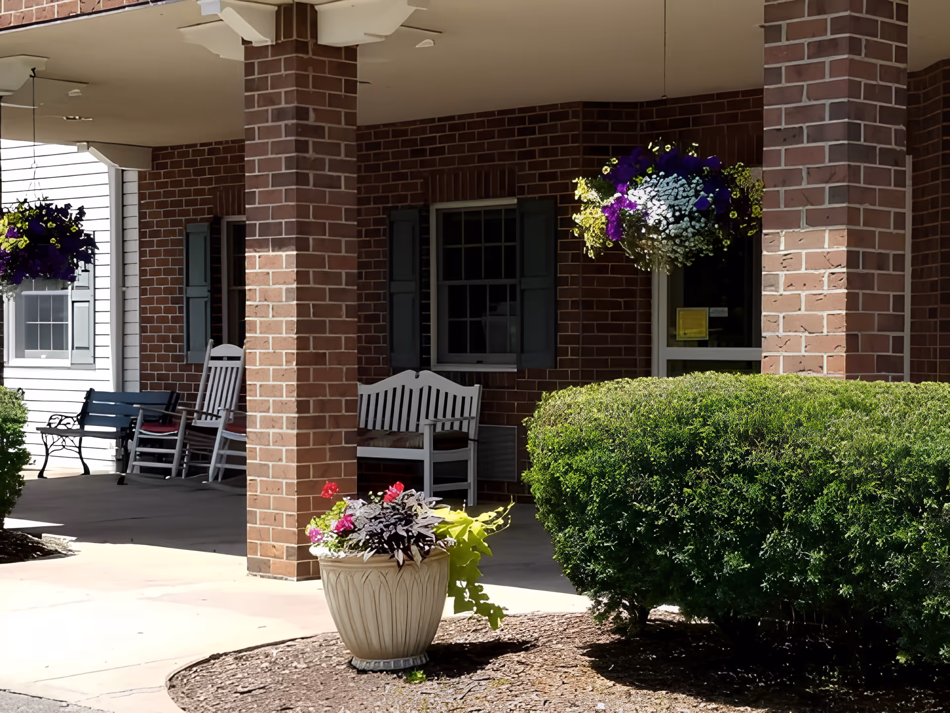 Covered outdoor patio area with brick pillars, white wooden benches, rocking chairs, hanging flower baskets with purple and white flowers, a large green bush, and a decorative planter with various plants and flowers.