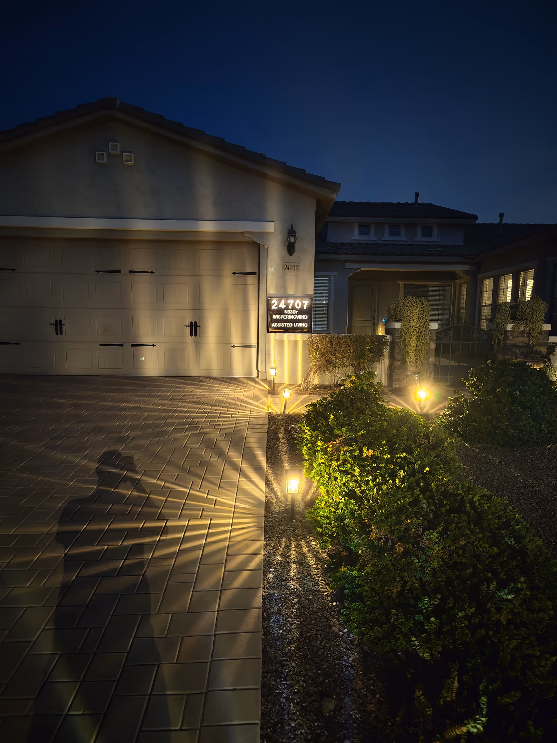 Night exterior of Whispering Wind Assisted Living with illuminated walkway and shrubs.
