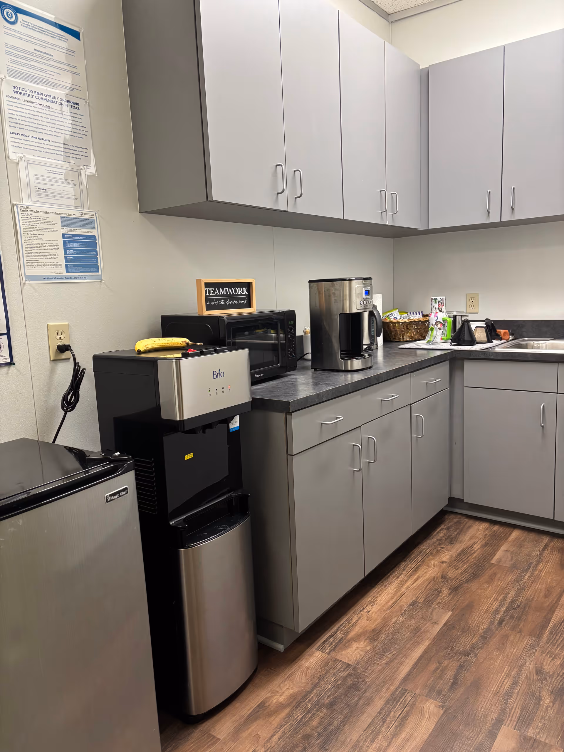 Grey cabinets line the walls with a coffee station and appliances in a kitchenette.