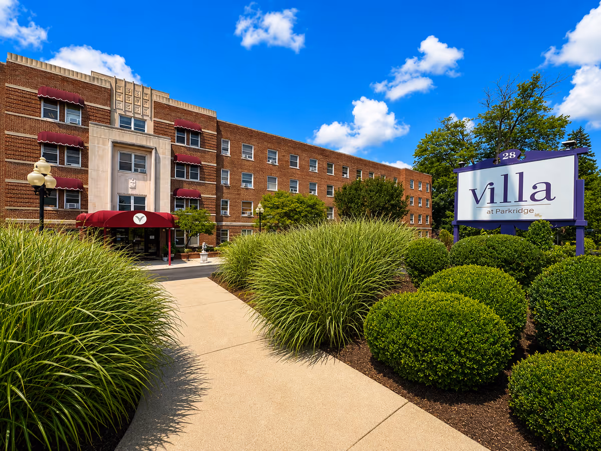 Brick senior living facility exterior with red awnings, manicured shrubs, and a Villa at Parkridge sign.