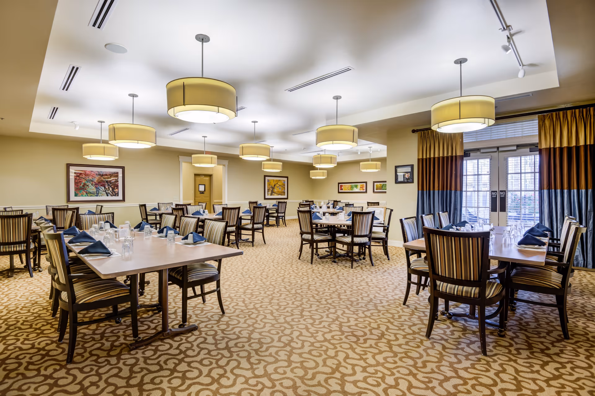 A spacious dining room with multiple rectangular tables set with glasses and folded napkins. The room features patterned carpet, striped upholstered chairs, large round ceiling lights, framed artwork on the walls, and large windows with curtains allowing natural light to enter.