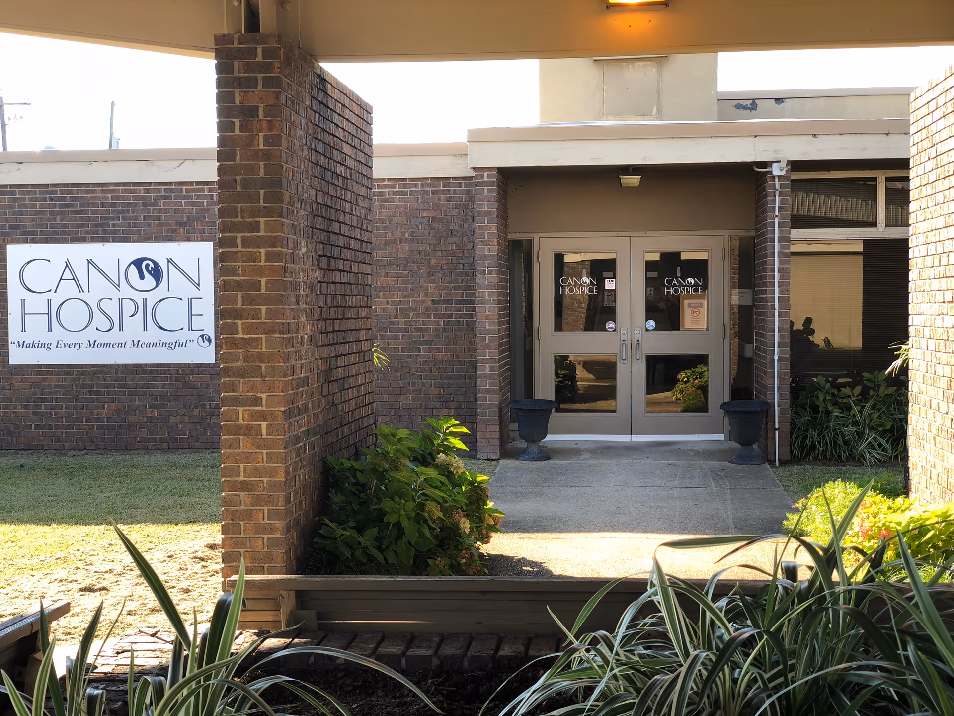 Brick building entrance with double doors labeled Canon Hospice, surrounded by plants.