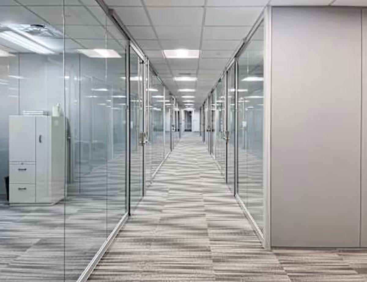 A glass-walled office hallway with striped carpet and ceiling tiles.