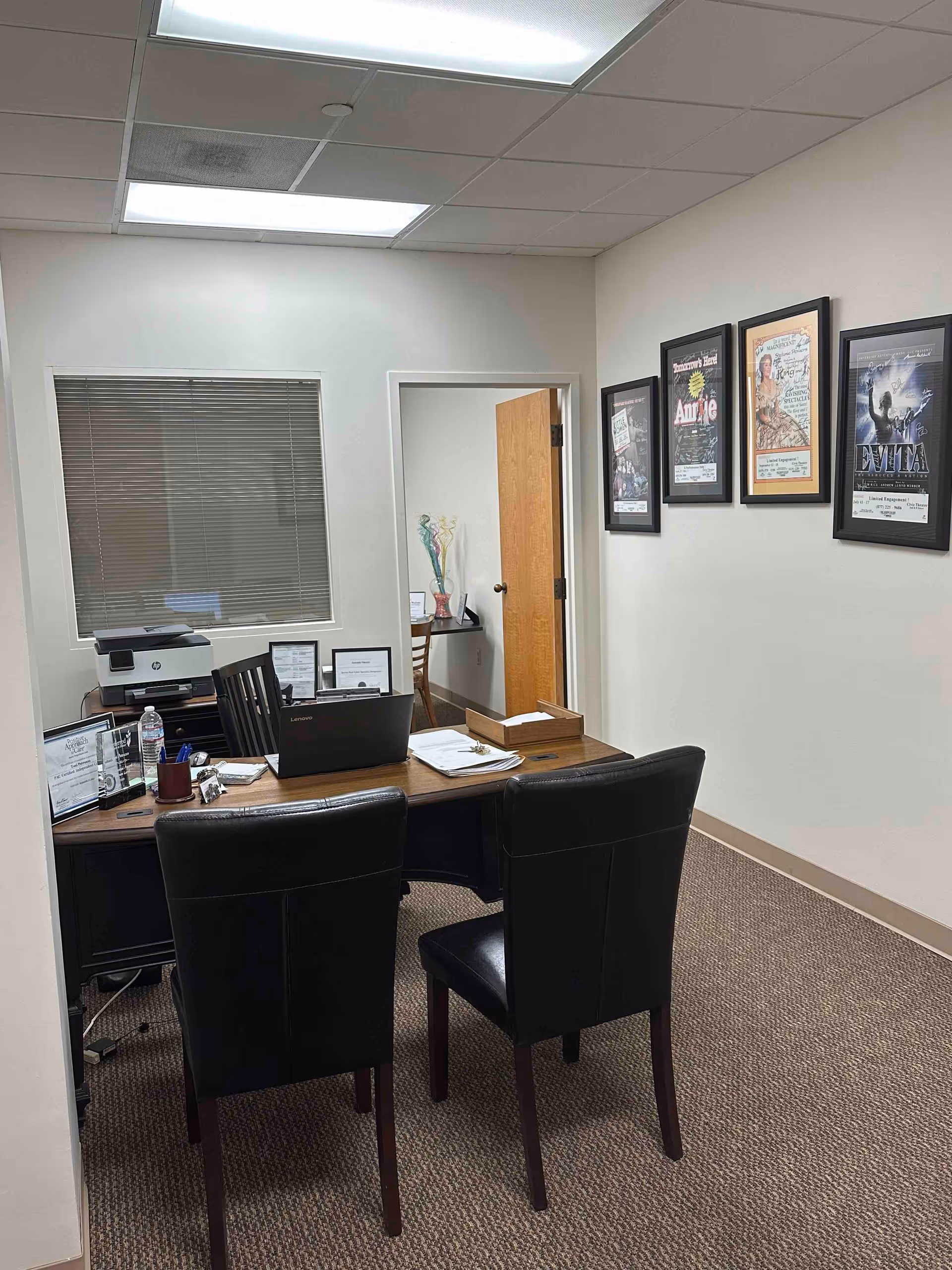 Office desk with two chairs, computer, printer, and framed posters on wall.