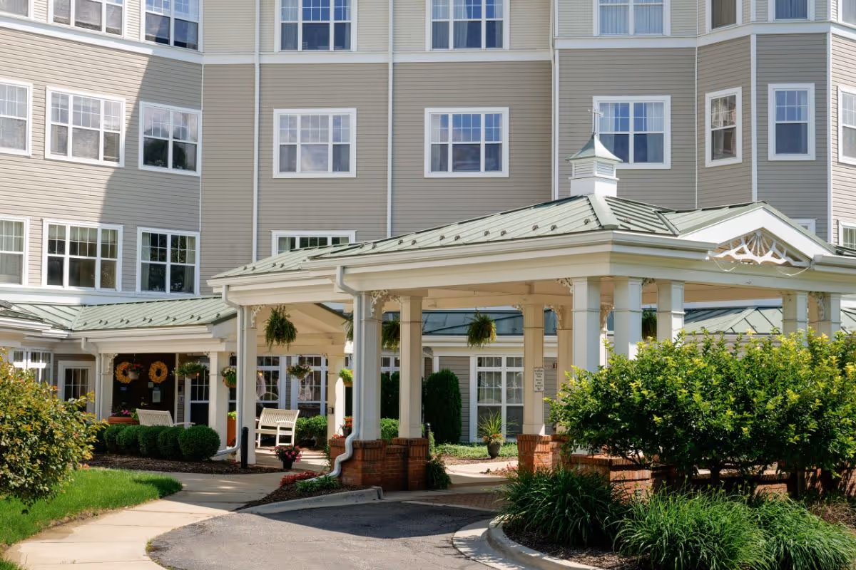 Covered entrance and landscaped front of a multi-story assisted living building with white columns and a green metal roof.