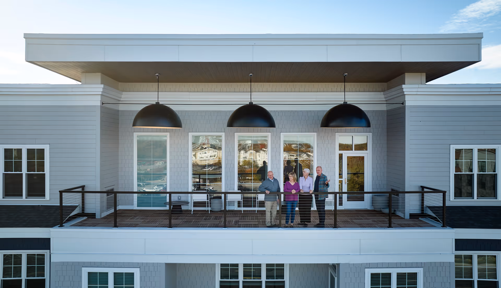 Four elderly people standing and talking on a balcony of a modern senior living facility building with large windows and three large black pendant lights hanging from the ceiling above the balcony.