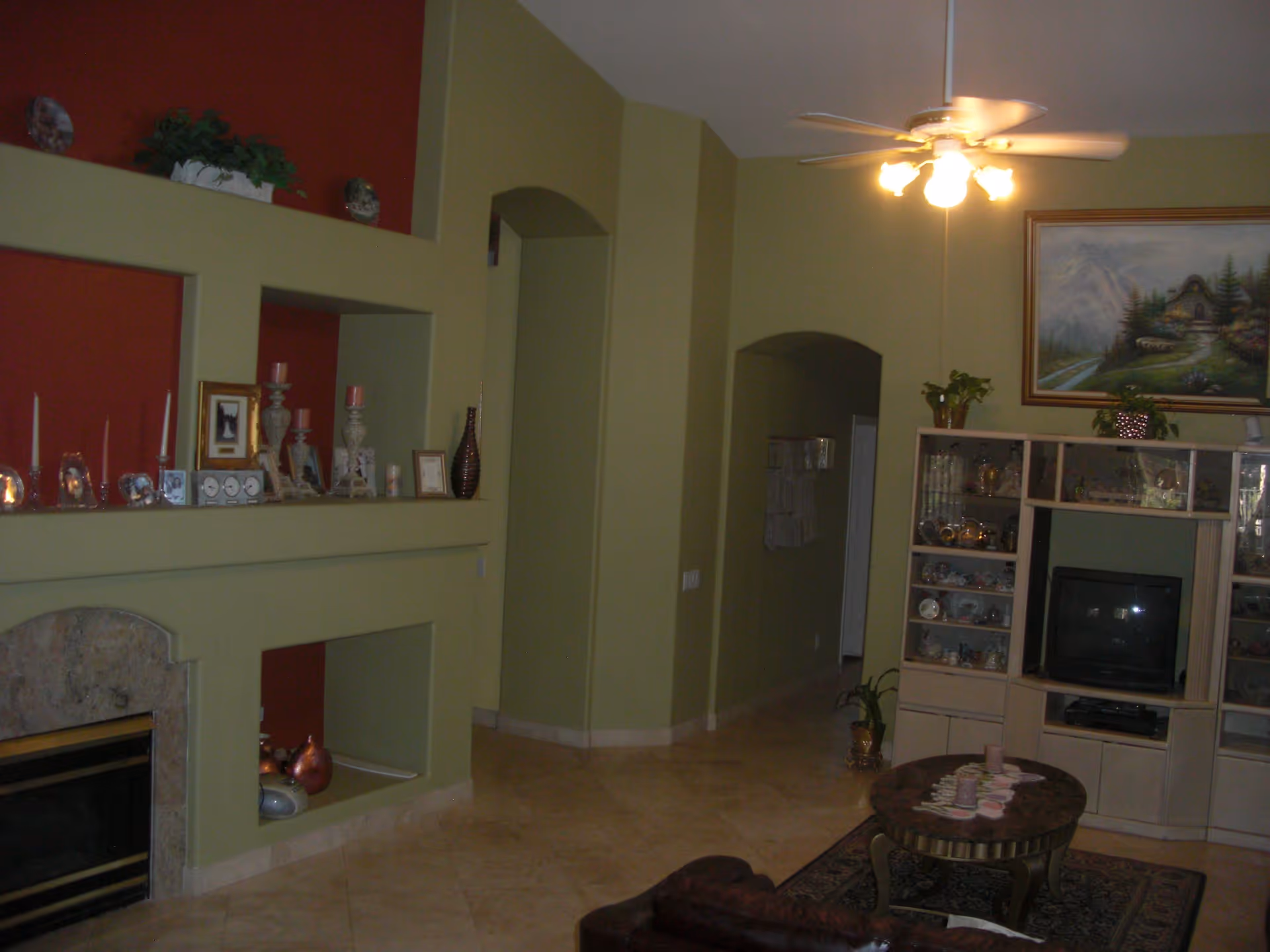 A living room with green walls and a ceiling fan with lights. There is a built-in fireplace with decorative items on the mantel, a wooden entertainment center with a TV and various collectibles, a round coffee table with coasters, and a leather chair partially visible. A painting of a mountain scene hangs above the entertainment center.