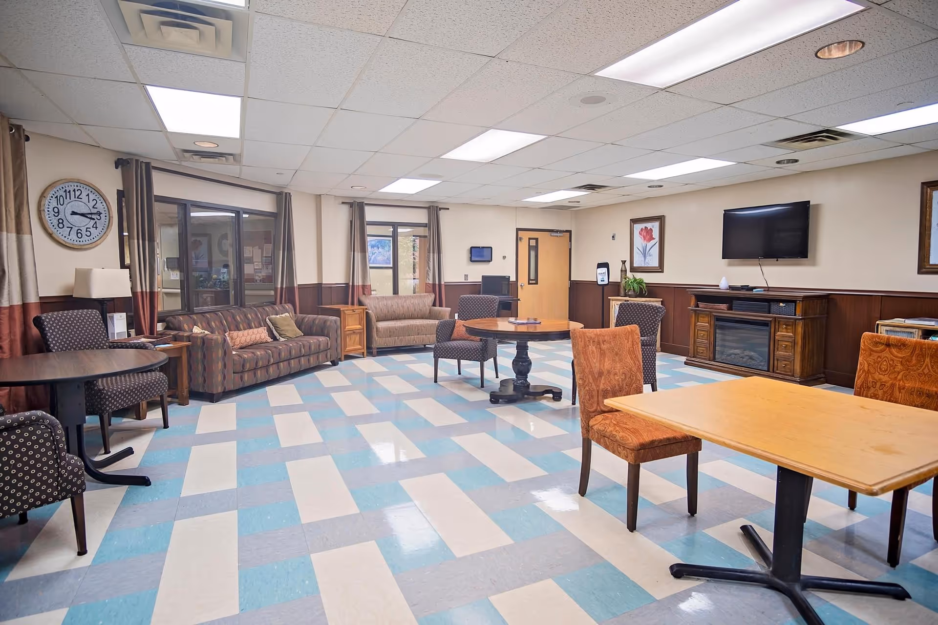 A spacious common area in a senior living facility with patterned blue and beige tiled flooring, multiple seating options including sofas and chairs, several tables, a wall-mounted TV above a wooden cabinet, framed artwork on the walls, and large windows with curtains allowing natural light.