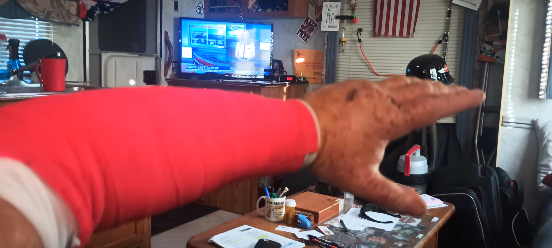 Cluttered living room scene with TV, papers, mug, and extended pink-sleeved arm in foreground.