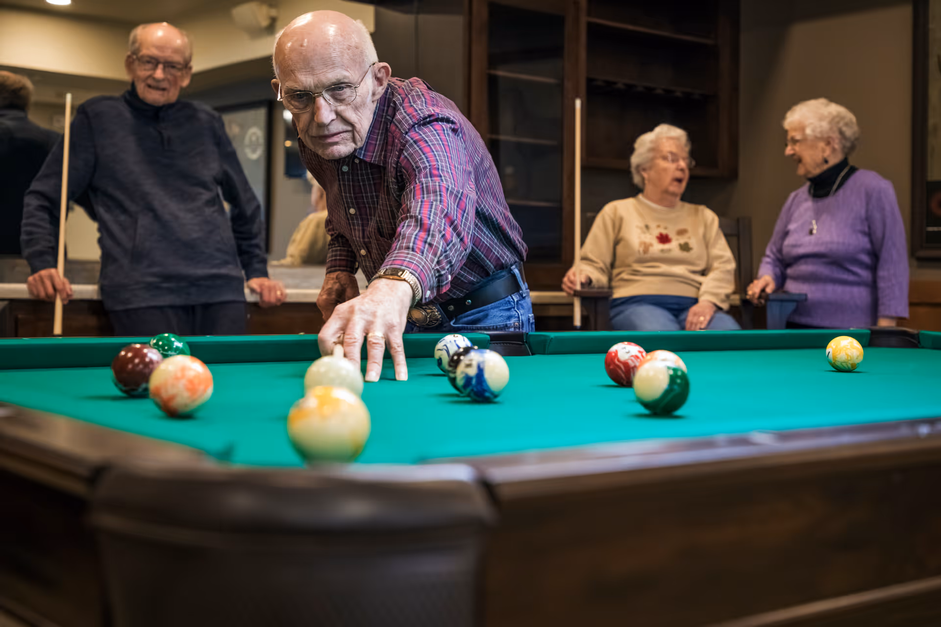 An elderly man in a plaid shirt is playing pool, aiming to hit the cue ball on a green pool table. Another elderly man stands behind him holding a pool cue, while two elderly women sit and chat in the background in a cozy indoor setting.