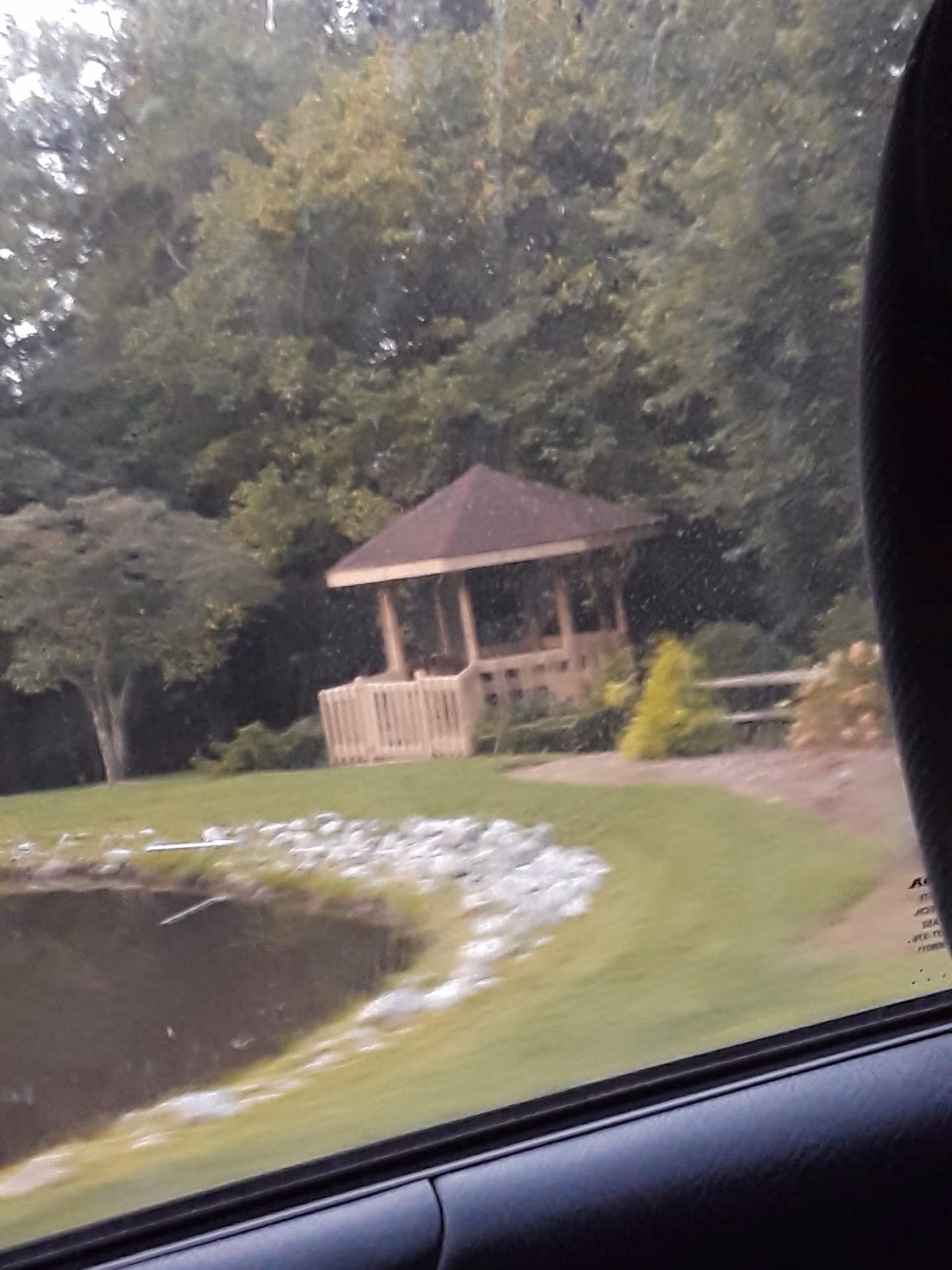 Wooden gazebo sits beside a grassy pond in a landscaped outdoor area.