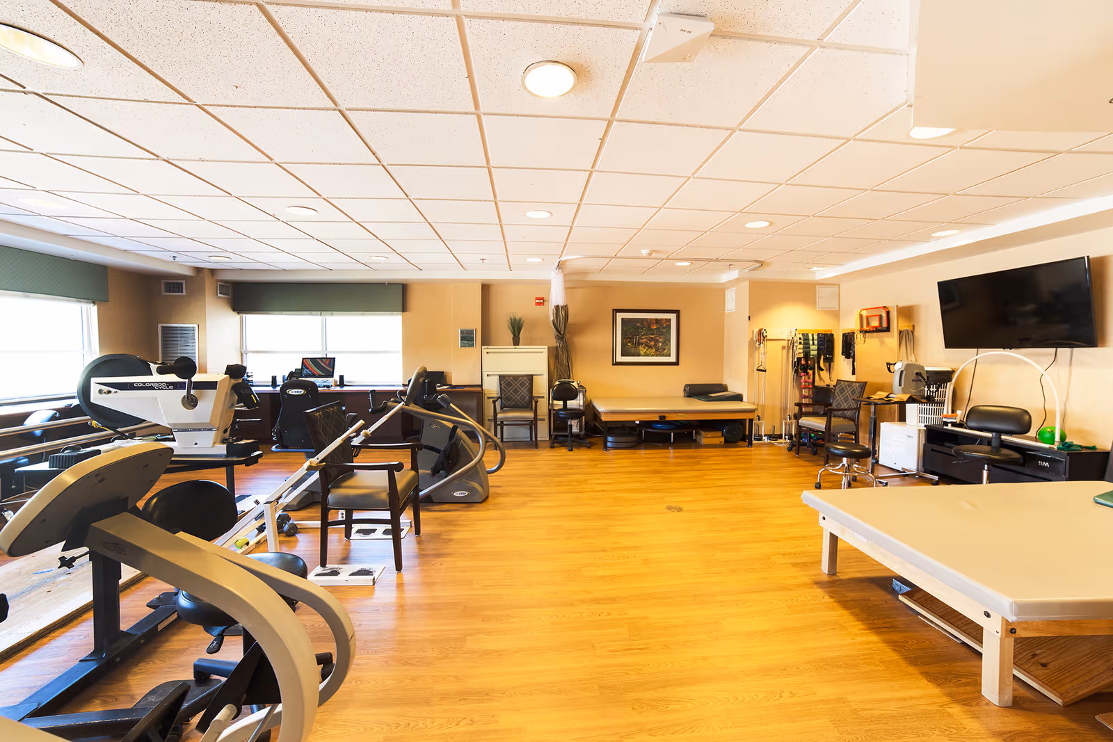 A spacious therapy or rehabilitation room with exercise equipment including stationary bikes, chairs, and therapy tables. The room has wooden flooring, a large wall-mounted TV, and windows with green valances allowing natural light to enter. Various therapy tools and supplies are organized along the walls.