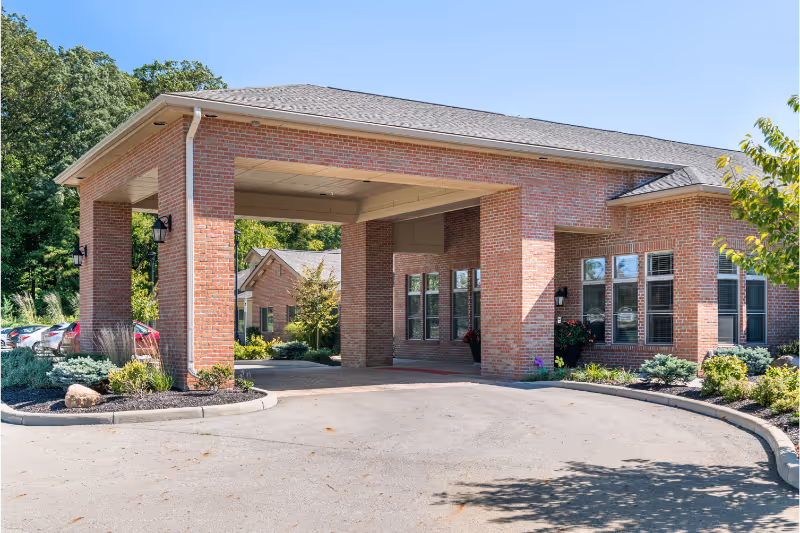 Exterior view of a brick building with a covered entrance driveway, surrounded by landscaped greenery and trees under a clear blue sky.