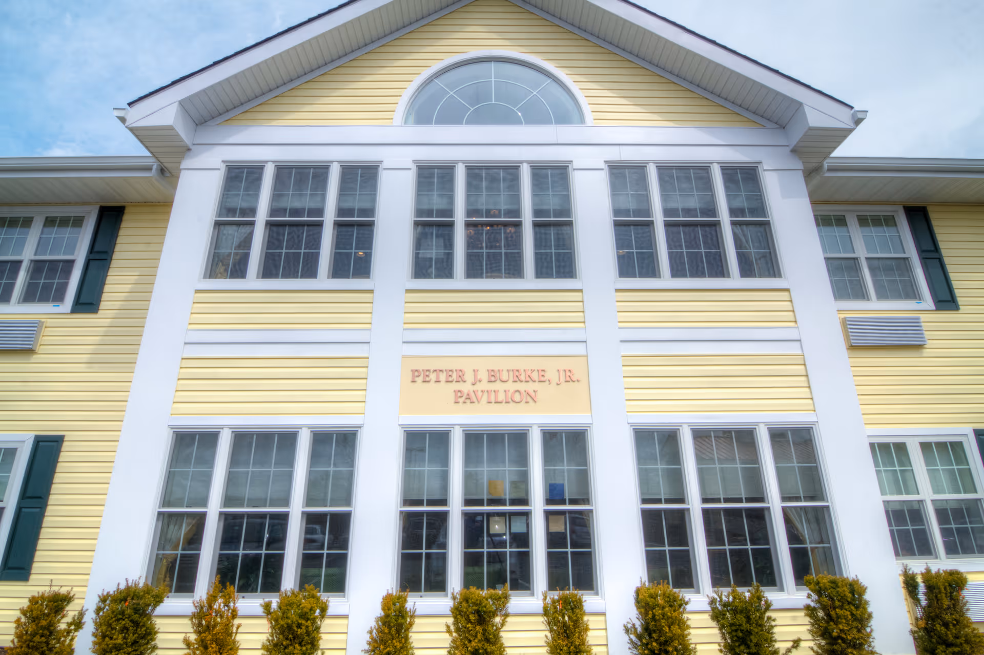 Front facade of a yellow two-story building with large windows and a sign reading "PETER J. BURKE, JR. PAVILION".