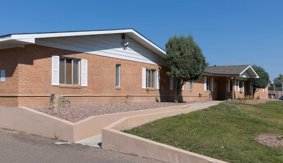 Low single-story brick senior living facility building with a front lawn, walkway, and trimmed trees under a clear blue sky.