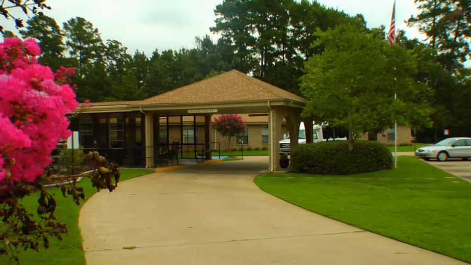 Entrance driveway leading to a covered drop-off area of a single-story nursing center building surrounded by green lawns, trees, and pink flowering bushes, with a few parked cars visible in the background.