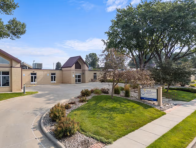 Exterior view of Good Samaritan Society - Luther Manor building with a driveway, landscaped greenery, trees, and a sign indicating the main entrance under a partly cloudy blue sky.