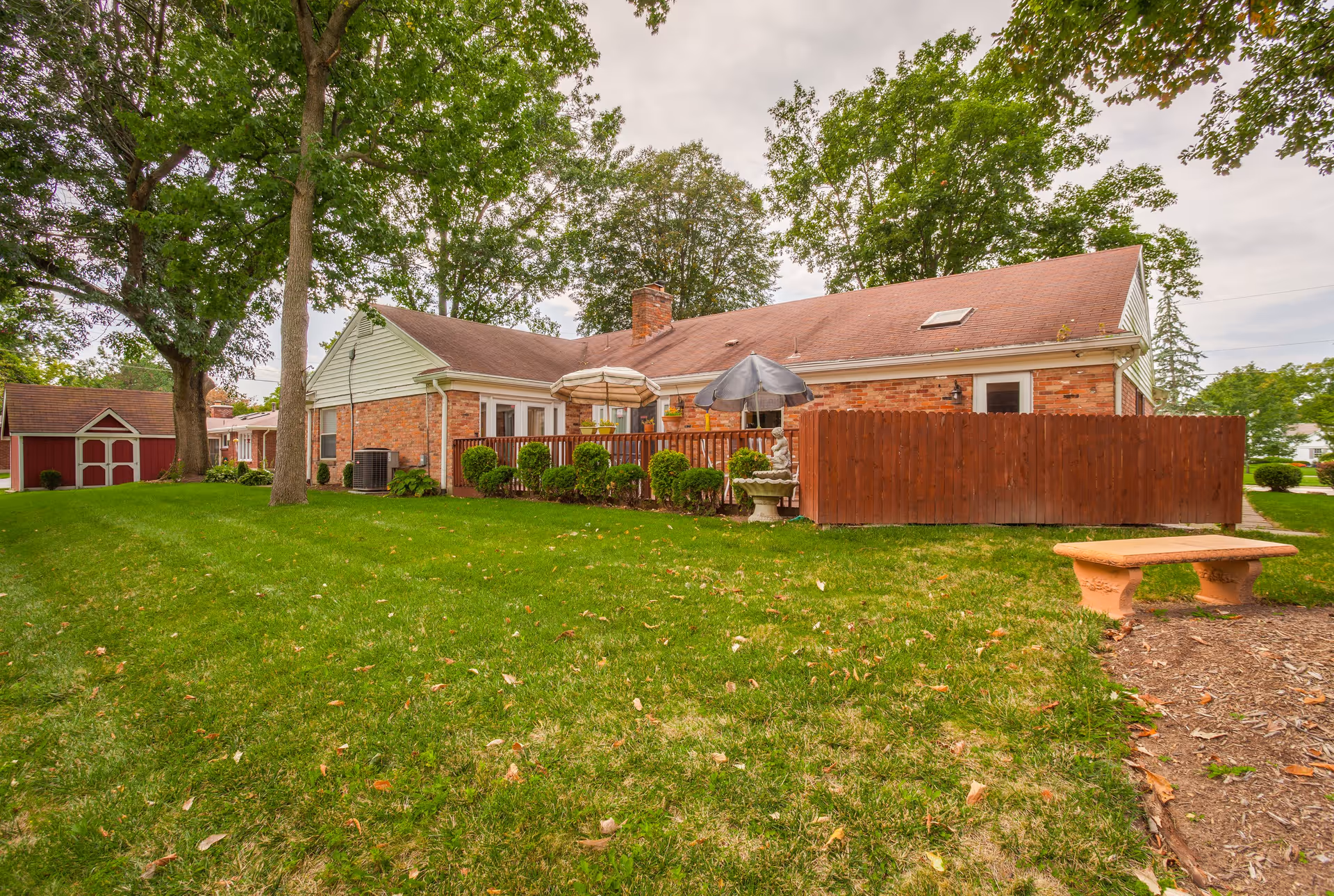 Single-story brick building with a fenced patio, umbrellas, green lawn, trees and a nearby bench.