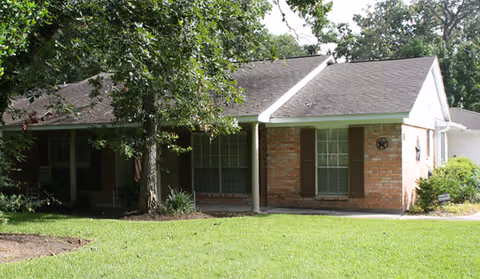 Single-story brick cottage with shaded front porch and green lawn in a residential setting