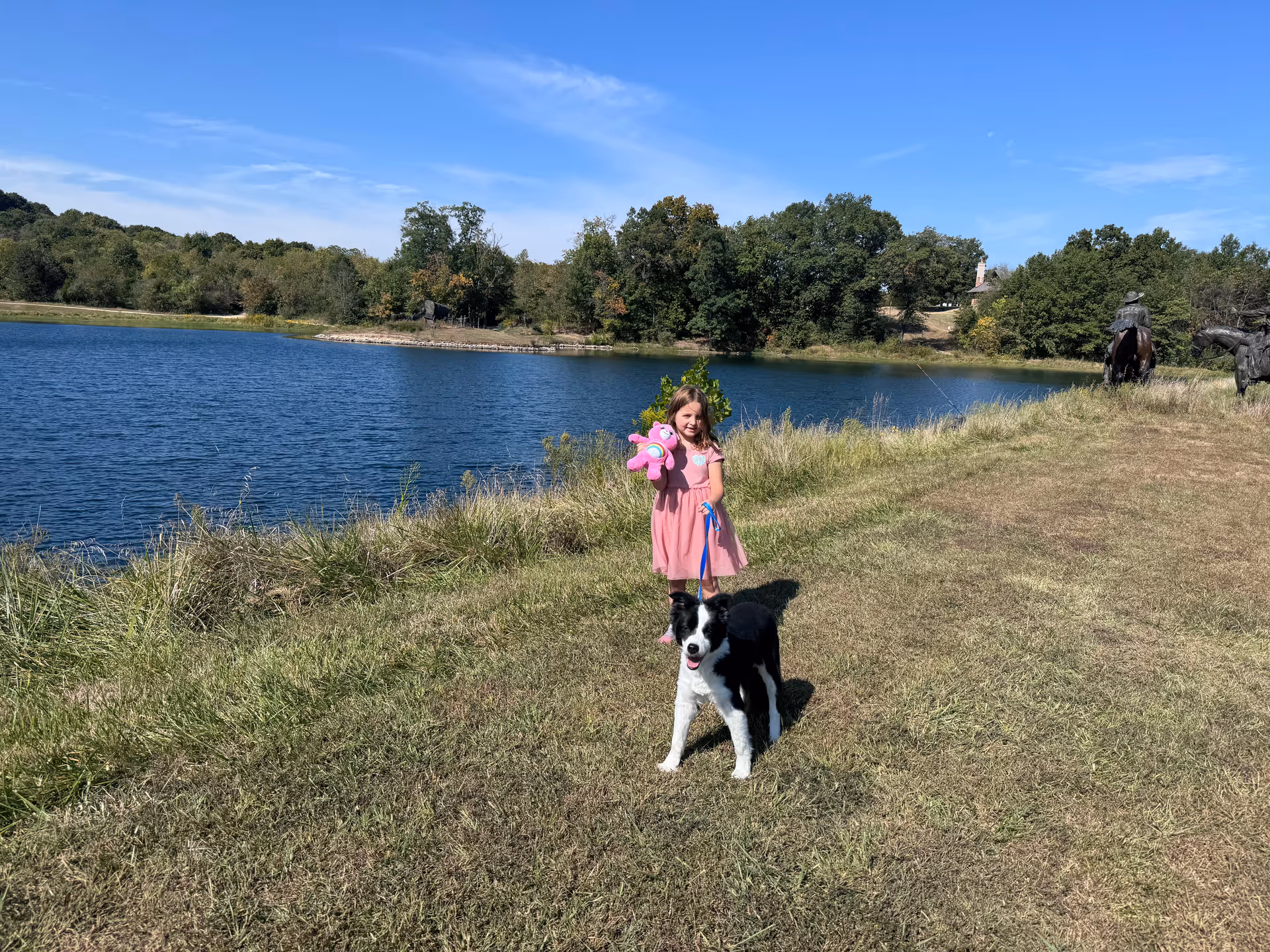 Young girl in a pink dress holds a plush toy and a dog beside a lake.