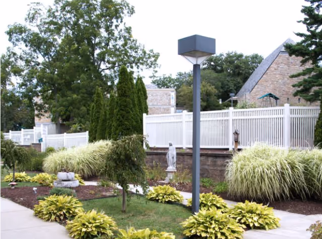 Outdoor garden area with green shrubs, small trees, and a statue. A white fence runs along the background with buildings and tall trees behind it. A modern square lamp post is visible near the center of the image.