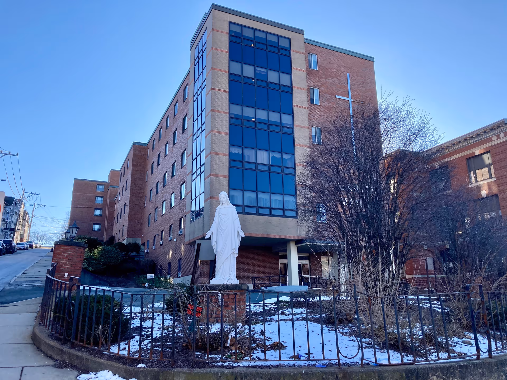Front exterior of a multi-story brick nursing home with a statue of the Virgin Mary in a fenced front garden.